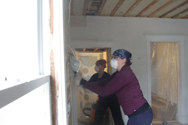 Photo of two women wearing dust masks and using crowbars to remove a plaster wall.